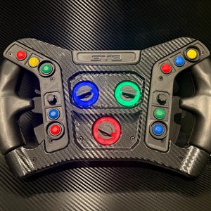 May include: A black carbon fiber racing steering wheel controller featuring the text GT3 embossed at the top center. The device includes multiple colorful push buttons in red, yellow, blue, and green, along with three large rotary dials in blue, green, and red.