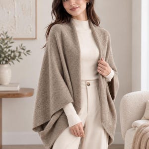 Puede incluir: Un poncho de punto beige abierto por delante con acabado texturizado, usado sobre una blusa de manga larga color crema con cuello alto y pantalones claros que presentan un botón visible en la cintura y un corte recto.