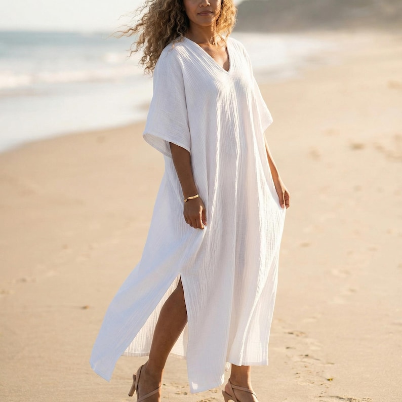 May include: A white maxi dress with a V-neck, short sleeves, and a high side slit. The dress is made of a lightweight fabric. The image shows the dress on a sandy beach. A gold bracelet is visible.