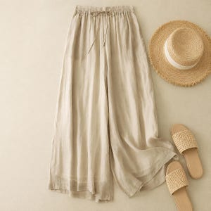 Peut inclure: Pantalon large en lin beige avec taille élastique et cordon de serrage, associé à un chapeau de paille et des sandales tressées. Le pantalon a une coupe décontractée, idéale pour le temps chaud. Le chapeau a un ruban blanc.