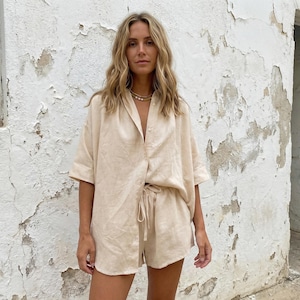 May include: A beige linen shirt and shorts set. The short-sleeved button-down shirt is paired with drawstring shorts. The outfit is made of a lightweight fabric, perfect for warm weather. The model is wearing a gold necklace.