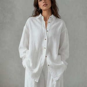 May include: A white linen shirt with a classic collar and button-down front, paired with matching white pants. The outfit has a relaxed fit and is made of a lightweight fabric. The shirt has long sleeves and wooden buttons.