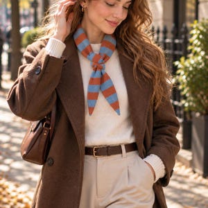May include: A brown wool coat worn over a white knit sweater and beige trousers with a brown leather belt. A blue and orange striped neck scarf is tied at the neck. A brown leather shoulder bag is carried on the side of the brown coat.