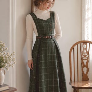 Puede incluir: Un vestido tipo pichi a cuadros de color verde oscuro con escote cuadrado, dos botones marrones en el corpiño y un cinturón marrón en la cintura. El vestido se lleva sobre un suéter de cuello alto acanalado de color crema y manga larga.