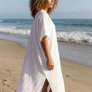 May include: A white beach cover-up dress with short sleeves and a side slit. The dress is made of a textured fabric. The image shows the dress on a sandy beach with the sea in the background. The dress is a plain white colour.