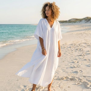 May include: A white, V-neck caftan dress with short sleeves. The dress is made of a lightweight fabric and has a relaxed fit. The model is standing on a sandy beach with the sea in the background. The dress is ankle-length.