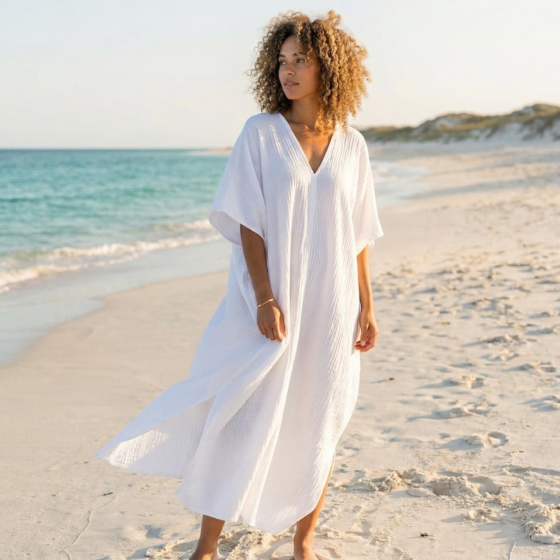 May include: A white, V-neck caftan dress with short sleeves. The dress is made of a lightweight fabric and has a relaxed fit. The model is standing on a sandy beach with the sea in the background. The dress is ankle-length.