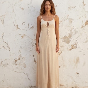 Könnte beinhalten: Ein hellbeige Maxikleid mit dünnen Spaghettiträgern und einem Schlüssellochdetail am Ausschnitt. Das Kleid hat eine fließende Silhouette und ist aus einem weichen, leichten Stoff gefertigt. Das Model steht vor einer strukturierten weißen Wand.