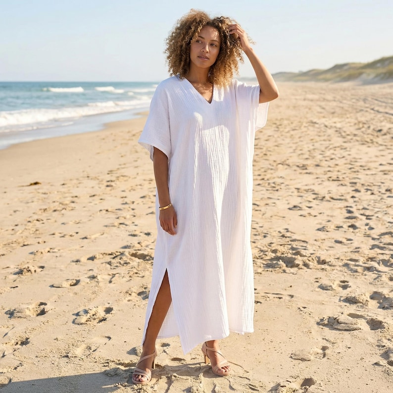 May include: A white, V-neck beach dress with short sleeves and a side slit. The dress is made of a textured fabric and is worn on a sandy beach with the ocean in the background. A gold bracelet is visible.