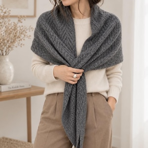May include: A charcoal grey triangular knit shawl featuring a textured chevron pattern, draped over a cream-coloured long-sleeve sweater and tan trousers. The shawl has a soft, fuzzy appearance and is wrapped around the shoulders and chest.