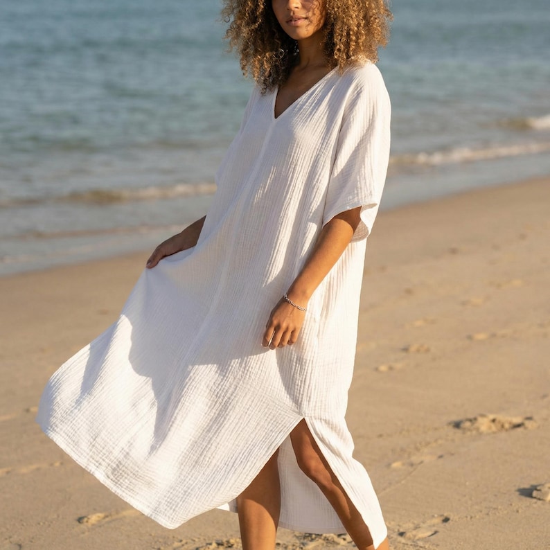May include: A white, V-neck dress with short sleeves and a side slit. The dress is made of a textured fabric and is knee-length. The model is standing on a sandy beach near the sea.