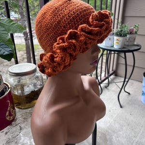 Può includere: Un cappello a secchiello all'uncinetto color arancione bruciato con un motivo a maglia denso e strutturato sulla calotta e un'ampia tesa arricciata che crea un effetto ondulato e stratificato sul bordo. Il cappello è in filato e forma rotonda.