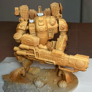 Pode incluir: Uma figura de ação robótica de cor bege sobre uma base texturizada com pequenos detalhes rochosos. A figura apresenta um grande canhão montado, lançadores de mísseis nos ombros e uma armadura mecânica com pequenos detalhes brancos na cabeça.