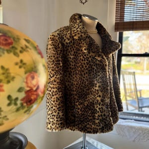 May include: A leopard print coat with a wide collar and button closure. The coat is displayed on a mannequin. The coat's pattern features brown spots on a tan background. A pearl necklace is visible on the mannequin.