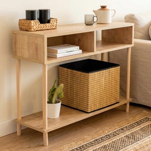 May include: A light wood-toned console table with two shelves and a lower shelf. The top shelf holds black canisters, a woven tray, a teapot, and a glass. A woven basket and a potted cactus are on the lower shelf.