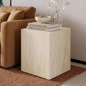 Peut inclure: Une table d'appoint carrée en pierre claire avec une pile de livres, un bol blanc et un vase en verre avec des tiges décoratives. Les livres indiquent "THE ART OF NATURAL STONE" et "MINIMALIST HOME DESIGN". Un canapé et un tapis marron sont en arrière-plan.