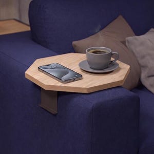 May include: A wooden sofa arm tray with a metal bracket, holding a smartphone and a cup of coffee on a saucer. The tray is light brown, and the coffee cup is gray. The sofa is dark blue, with gray and brown pillows.