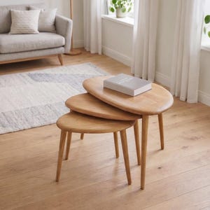 Puede incluir: Un juego de tres mesas nido de madera clara con forma ovalada. Las mesas tienen patas delgadas y cónicas. Un libro descansa sobre la mesa superior. El conjunto está en un salón con un sofá y una alfombra.