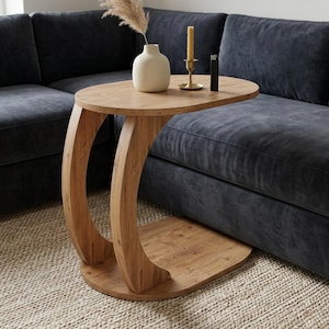 May include: A light brown wooden side table with a curved base and oval top. A white vase with dried pampas grass, a candle, and a lighter sit on the table. The table is next to a dark blue sofa. The table's height is approximately 60 cm.