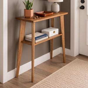 May include: A light-brown wooden console table with a lower shelf and angled legs. The top shelf holds a small lamp, a leather tray, and a potted plant. A white marble box and books are on the lower shelf. The table is approximately 100 cm in length.