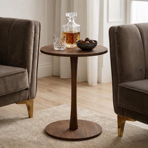 May include: A dark brown, round side table with a single stem and circular base. A crystal decanter, a rocks glass, and a small bowl of nuts sit on top. Two brown armchairs with gold legs are on either side.