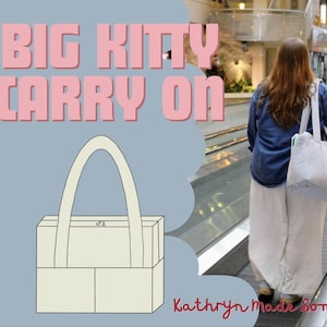May include: A light blue tote bag with white vertical stripes is carried by a person on an escalator. The bag has a large, arched handle. The words "BIG KITTY CARRY ON" are in pink block letters. The text "Kathryn Made Something" is in cursive.
