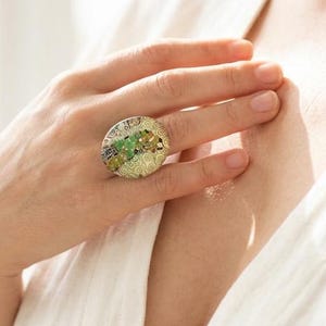 May include: A large, circular statement ring featuring a textured, metallic gold-tone base with an intricate, raised pattern. The center is embellished with a cluster of small, multi-colored gemstones in shades of green, yellow, and brown.