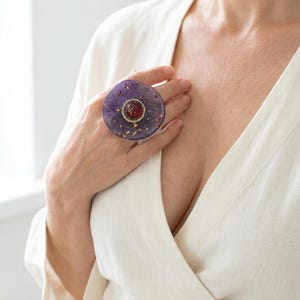 Puede incluir: Un anillo grande y redondo con una base de resina morada, copos dorados y una piedra roja central. El anillo está engarzado en un metal dorado y se lleva en una mano.