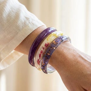 Puede incluir: Cuatro brazaletes de resina en tonos morado, blanco y dorado apilados en una muñeca. Los brazaletes presentan detalles florales y de pan de oro incrustados. La muñeca está al lado de una camisa blanca de manga larga.