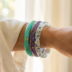 May include: Three colorful resin bangles are stacked on a wrist. The bangles are turquoise, purple, and clear with gold flecks. The clear bangle has blue flecks. The wrist is next to a white linen sleeve.
