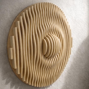 Könnte beinhalten: Runde Wandkunst aus hellem Holz mit einem geschichteten, wellenartigen Design. Das helle Holz ist in vertikalen, wellenförmigen Linien angeordnet, wodurch ein dreidimensionaler Effekt entsteht. Ein zentrales, kugelförmiges Element verleiht dem Kunstwerk Tiefe.