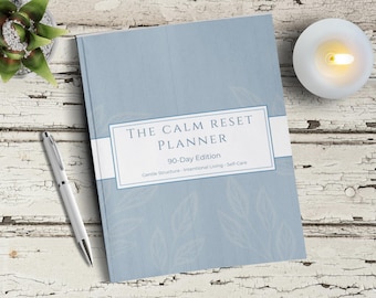 Puede incluir: Un planificador azul claro "The Calm Reset Planner" con una etiqueta blanca, un bolígrafo plateado, una pequeña suculenta y una vela encendida. El planificador tiene el texto "90-Day Edition" y "Gentle Structure - Intentional Living - Self-Care".