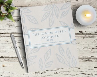 Puede incluir: Un diario titulado "The Calm Reset Journal" con un patrón de hojas azul claro. El diario tiene el texto "30 días" y "Un diario guiado para mujeres que buscan calma y estructura". Un bolígrafo plateado, una suculenta y una vela encendida también están en la imagen.