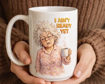 Puede incluir: Taza de cerámica blanca con una ilustración humorística de una anciana y el texto "I AIN'T READY YET". La mujer lleva gafas y una camisa de pijama rosa con flores, sosteniendo una taza de café. La taza tiene un asa blanca.