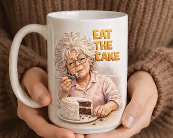 Puede incluir: Taza de cerámica blanca con una ilustración de una anciana comiendo pastel. La taza tiene el texto "EAT THE CAKE" en letras doradas. La mujer lleva gafas y una camisa rosa con flores.