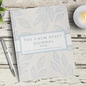May include: A journal titled "The Calm Reset Journal" with a light blue floral design. The journal includes the text "90-Day" and "A Guided Journal for Women Seeking Calm and Structure." A silver pen and a lit candle are also visible.