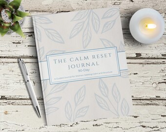Puede incluir: Un diario titulado "The Calm Reset Journal" con un diseño floral azul claro. El diario incluye el texto "90-Day" y "A Guided Journal for Women Seeking Calm and Structure". También se ven un bolígrafo plateado y una vela encendida.