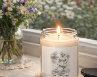 Puede incluir: Una vela encendida en un tarro de cristal con una etiqueta que muestra una ilustración en blanco y negro de una persona. La vela está sobre un posavasos de madera. Un jarrón con flores está en el fondo. La etiqueta dice "Pink State Designs."