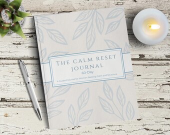 Puede incluir: Un diario titulado "The Calm Reset Journal" con un diseño floral azul claro, un bolígrafo plateado y una vela encendida sobre una superficie de madera blanca desgastada. El diario está etiquetado como "60 días" e incluye el texto "Un diario guiado para mujeres que buscan calma y estructura."