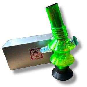 May include: A neon green water pipe with a black base and a clear glass mouthpiece. The pipe has a cylindrical chamber and a curved design. A silver box with a red and white design is next to the pipe.