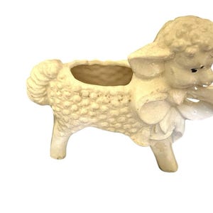 May include: A cream-colored ceramic planter shaped like a lamb with a textured, curly wool surface. The lamb features a large bow around its neck, a small opening on its back for plants, and dark painted eyes, standing on four short legs.