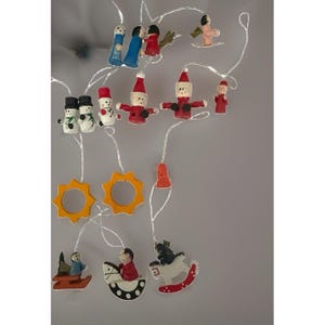 Puede incluir: Una guirnalda de adornos decorativos con pequeñas figuras de madera pintadas a mano, incluyendo muñecos de nieve, Papá Noel, ángeles y caballos balancín, unidos por cables blancos con pequeñas luces LED integradas.
