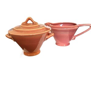 Puede incluir: Un juego de servir de cerámica vintage en tonos coral y rosa. El juego incluye un cuenco con tapa y asas y una jarra de crema con asa. Ambas piezas tienen un acabado brillante y detalles acanalados horizontales.