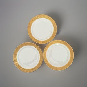 May include: Three circular white porcelain saucers arranged in a triangular formation. Each saucer features a wide, ornate gold rim decorated with a detailed, repeating embossed pattern along the outer edge and a smooth, plain white center.