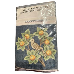 May include: A William Morris needlepoint miniature kit titled Woodpecker 2. The canvas features a bird perched among green leaves and orange fruit against a dark blue grid background, all sealed in a clear plastic protective sleeve.