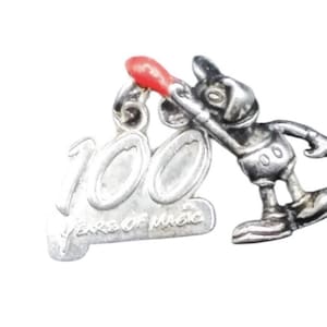 May include: A silver-toned metal charm featuring a cartoon mouse character leaning against the number 100. The number includes the text YEARS OF MAGIC and the character holds a small red-tipped matchstick. The charm has a small loop at the top.