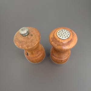 May include: Two wooden salt and pepper shakers with a natural brown finish. The left shaker features a single metal screw top, while the right shaker has a circular metal cap with multiple small perforations for dispensing seasoning.