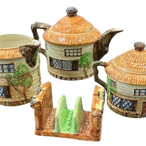 Puede incluir: Un juego de té de cerámica que incluye una tetera, una jarra de leche, un azucarero con tapa y un soporte para tostadas. Cada pieza está pintada como una cabaña con techo de paja, paredes de piedra, ventanas de celosía y árboles verdes.