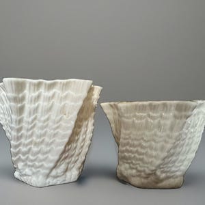 May include: Two white ceramic vessels with a distinct ruffled, shell-like texture. The taller vessel on the left features a bright white finish, while the shorter vessel on the right has a subtle greyish tint near the base and rim.
