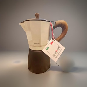 May include: A cream and dark brown moka pot with a wood-effect handle and lid knob. A square tag hangs from the handle by a red, white, and green string, featuring the text gnali & zani and an Italian flag graphic on a white background.